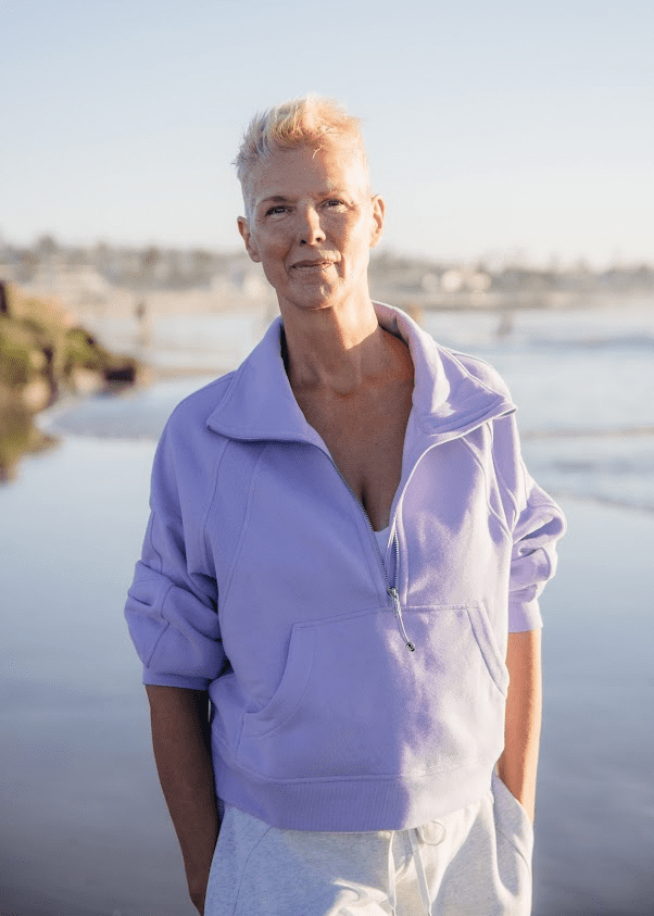 A woman in a light purple jacket standing near a beach on a sunny day.
